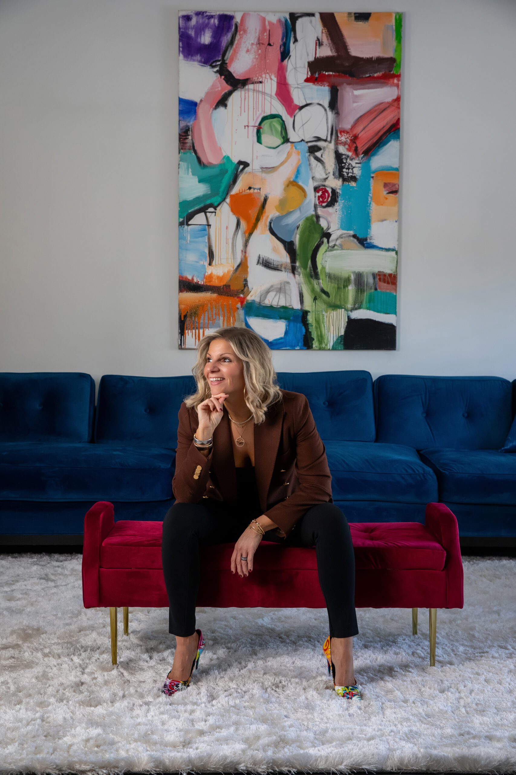 Woman seated on a red bench, wearing a brown blazer and black pants. She is in front of a blue sofa and an abstract colorful painting on a white wall.
