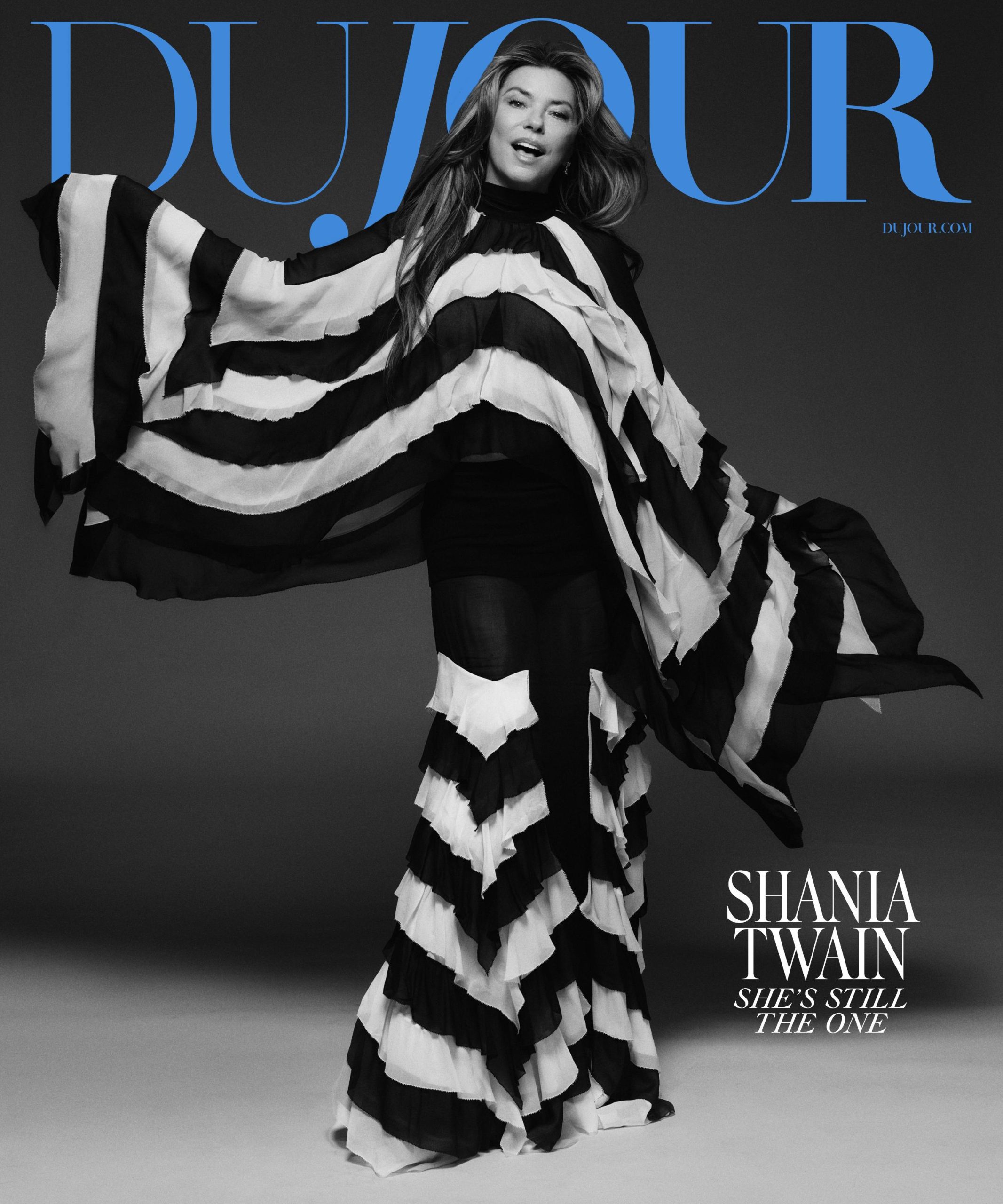 A woman poses in a flowing, striped black and white outfit against a plain backdrop on the cover of DuJour magazine with the headline "Shania Twain: She's Still The One.